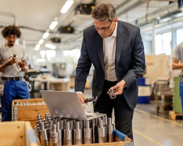 factory-manager-reading-instructions-computer-while-checking-AMICS-manufacturing-ERP-steel-bolt-industrial-building-manual-workers-are-background